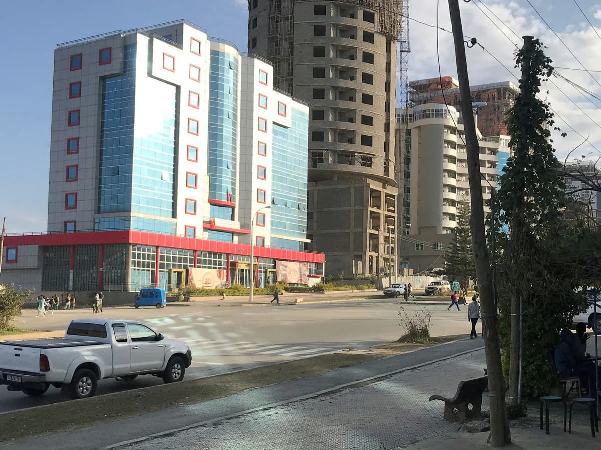 A street in Mekelle with mid-rise buildings, a bajaj and passing traffic.